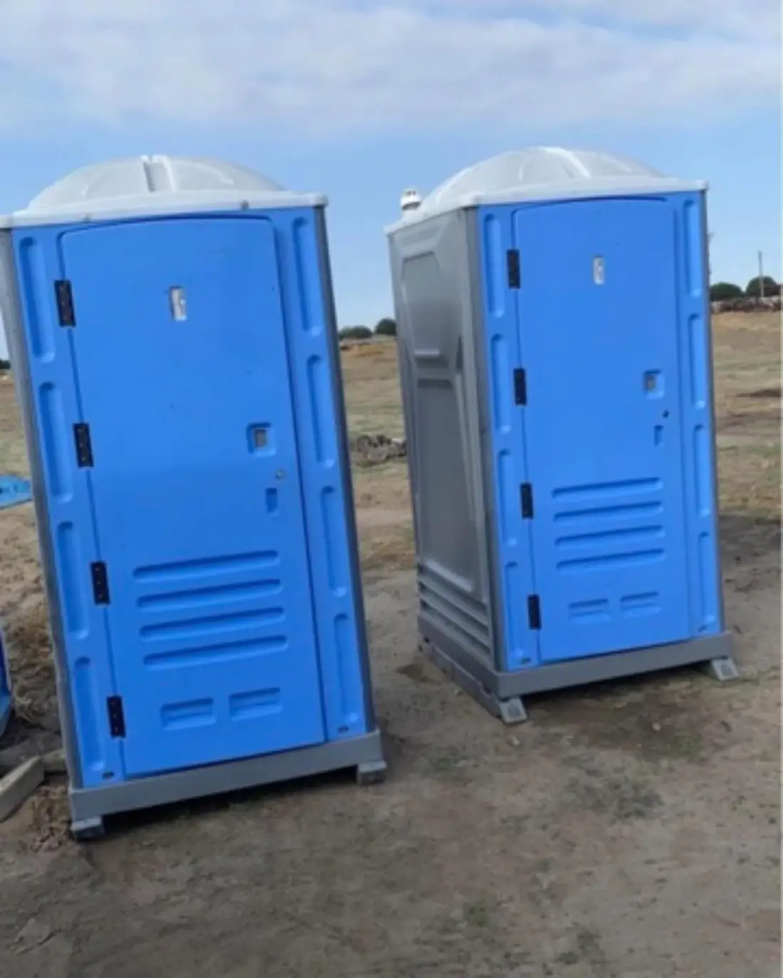 Two portable toilets, one blue and one gray, standing on an open dirt lot
